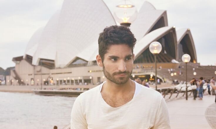 Ein Studennt mit dunklen Haaren und im weißen T-Shirt steht vor dem Sydney Opera House im Hafen Sydneys.
