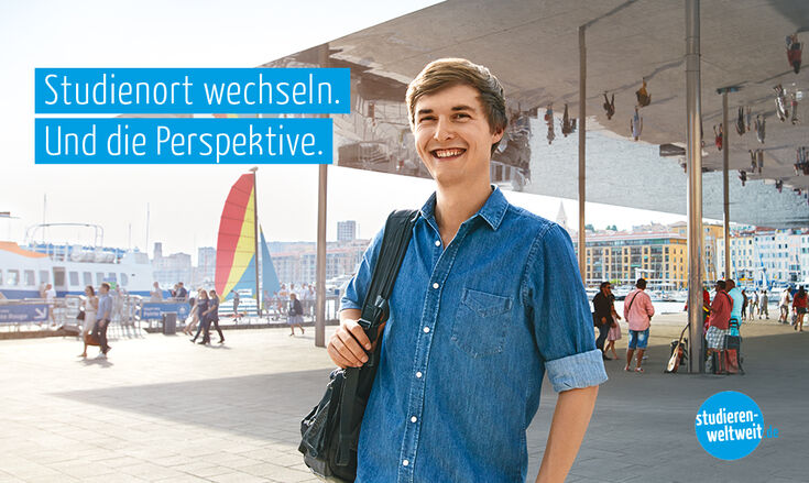Ein Student steht mit einem dunklen Rucksack über der Schulter am Hafen mit anlegenden Schiffen.