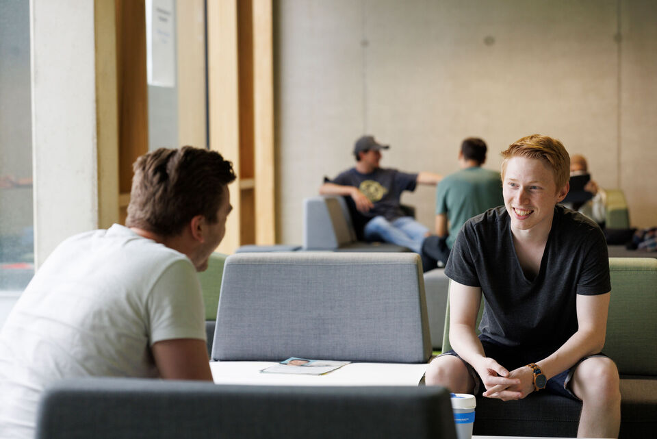 Two male students are sitting in one of the lounge areas in our J-building on campus Krefeld-South talking to each other. 