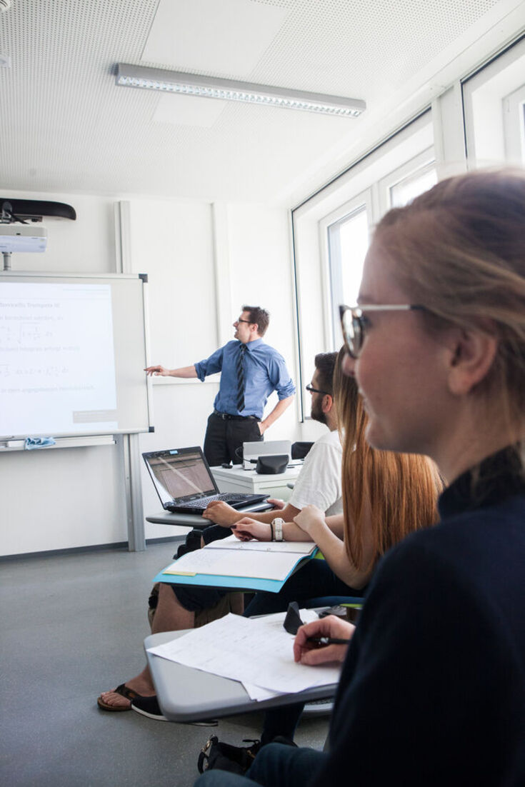 Studierende sitzen im Seminarraum; ein Dozent steht vor dem Whiteboard und zeigt darauf. 